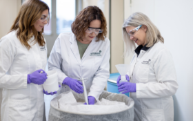 3 women in lab coats around a barrel